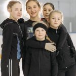 A group of young girls smiling and posing on an indoor ice rink wearing warm winter clothing.