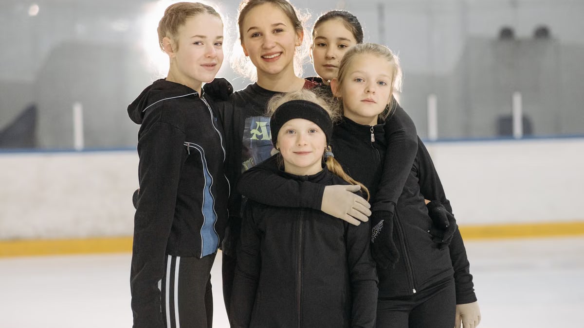 A group of young girls smiling and posing on an indoor ice rink wearing warm winter clothing.